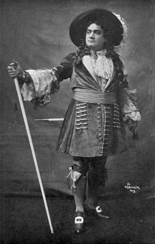 Fotografia: śpiewak w kostiumie z epoki, w kapeluszu z piórami i z laską, w teatralnej pozie